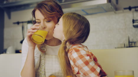 Attractive-mother-in-the-apron-drinking-juice-and-her-cute-daughter-kissing-her-in-the-cheek,-than-mom-and-daughter-kissing-and-hugging-in-the-kitchen.-Indoors